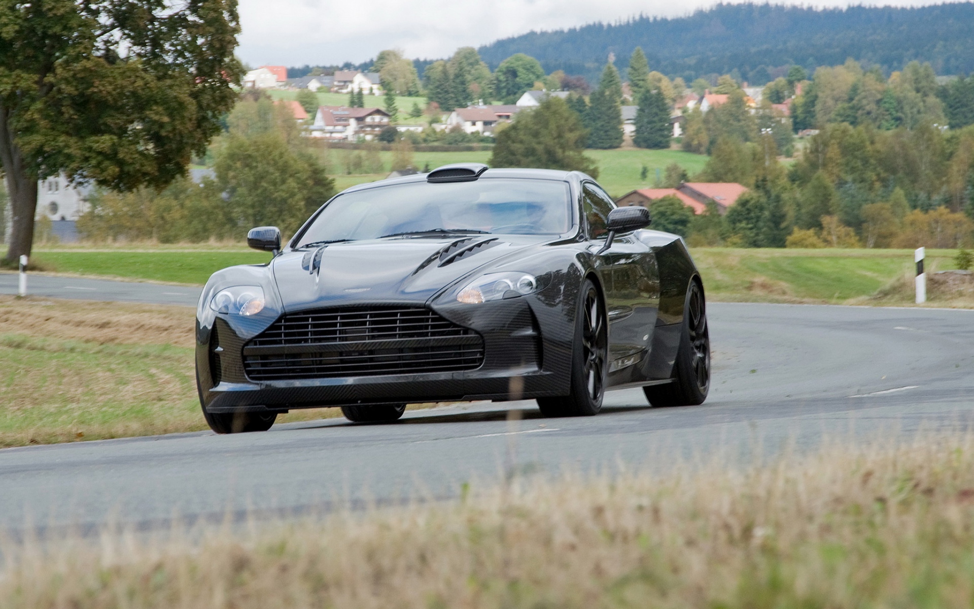 Картинки Mansory cyrus, 2009, черный, вид спереди, стиль, aston martin, дома, деревья, асфальт фото и обои на рабочий стол